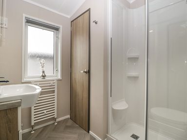A bathroom with a shower enclosure a sink a window with a vase of flowers and a wooden door at 14 Fairways in Burnham-On-Sea