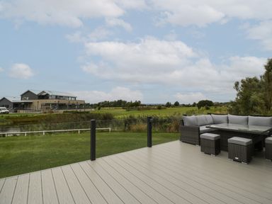 An outdoor deck with gray wicker corner sofa and stools overlooking a grassy golf course and a clubhouse at 14 Fairways in Burnham-On-Sea
