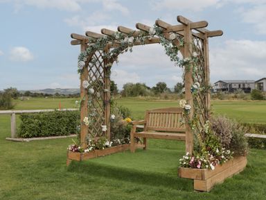 A wooden bench under a wooden pergola decorated with flowers and greenery on a grass lawn at 14 Fairways in Burnham-On-Sea