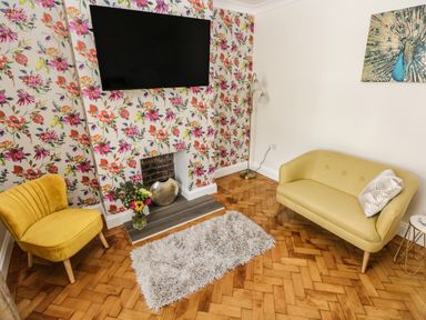 A sitting room with floral wallpaper and a wall-mounted TV a yellow armchair a yellow sofa a floor lamp a small rug on a wooden floor and a peacock painting at Larkhill in Tenby