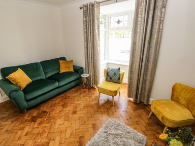 A living room with a green sofa, two yellow chairs, a small round side table, curtains, a window with patterned glass, and a rug on a wooden floor at Larkhill in Tenby