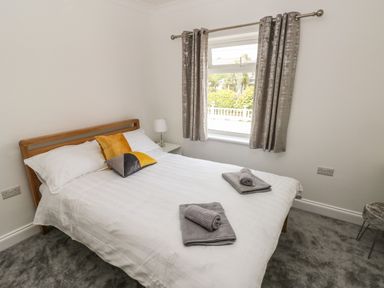 A bedroom with a double bed white bedding two gray towels on the bed bedside table with lamp and window with curtains at Larkhill Tenby