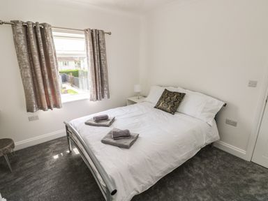 A bedroom with a double bed with white bedding and gray towels on top a window with curtains and a bedside table with a lamp at Larkhill in Tenby