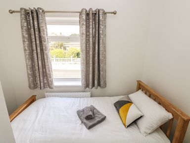 A bedroom with a wooden bed frame white bedding two pillows and folded towels on the bed in front of a window with patterned curtains at Larkhill in Tenby