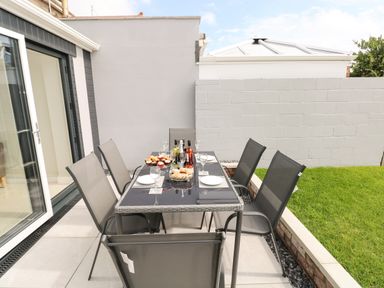 An outdoor patio dining table set with six chairs plates glasses and bottles near a grass lawn at Larkhill in Tenby