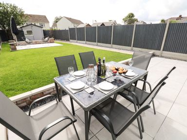 An outdoor patio with a glass table and six chairs set for dining next to a lawn and a shed with two chairs at Larkhill in Tenby
