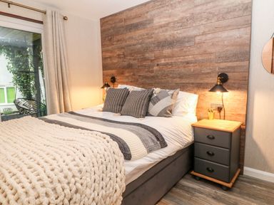 A bedroom with a bed, bedside table, wooden paneled wall, and window with curtains at Lavender Lodge in St Ann's Chapel Cornwall