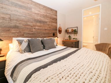 A bedroom with a bed with multiple cushions a chunky knit blanket a wooden accent wall a chest of drawers with plants and shelves a brown chair and an open door revealing a bathroom at Lavender Lodge in St Ann's Chapel Cornwall