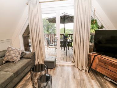 A living room with a sofa, two black metal wire tables, a television on a wooden stand, sliding glass doors with curtains leading to a balcony with outdoor furniture and an umbrella at Lavender Lodge in St Ann's Chapel Cornwall