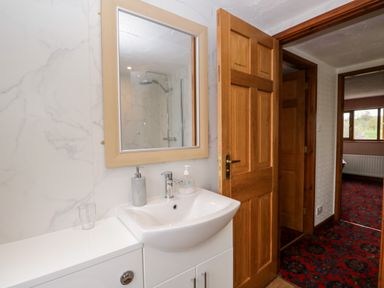 A bathroom sink with a mirror above and an open wooden door showing a hallway with a patterned red carpet at Rose Cottage in Burneside