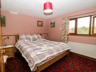 A bedroom with a double bed with plaid bedding bedside tables lamps paintings a window and floral curtains at Rose Cottage in Burneside