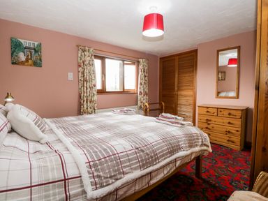 A bedroom with a bed covered in plaid bedding a wooden dresser with a mirror floral curtains and a red carpet at Rose Cottage in Burneside