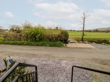 A view of a gravel driveway, steps with a black railing, trimmed hedges, a small paved area, and a green field with scattered sheep at Rose Cottage in Burneside