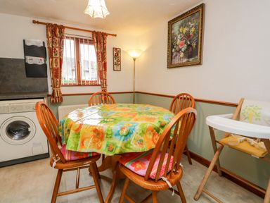 A dining area with a round table covered with a colorful plastic tablecloth surrounded by four wooden chairs and a high chair next to a window with patterned curtains at Rose Cottage in Burneside