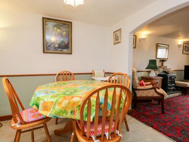 A dining area with a round table covered with a colorful tablecloth and wooden chairs next to a living room with armchairs and a fireplace at Rose Cottage in Burneside