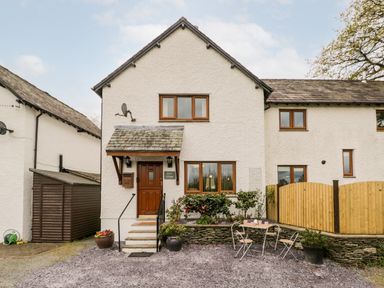 A white exterior of a cottage with wooden doors and windows a small seating area with a table and chairs outside and potted plants at Rose Cottage in Burneside