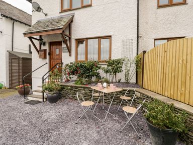 An outdoor patio area with a small table and three chairs in front of a white house with wooden doors and windows at Rose Cottage Burneside