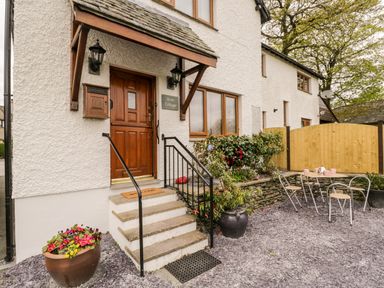 The entrance of a white cottage with wooden door and steps leading up to it with outdoor table and chairs nearby at Rose Cottage Burneside