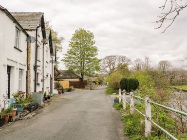 A country road with white houses on the left and a river with railings on the right at Rose Cottage in Burneside