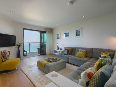 A living room with gray sectional sofa, yellow chair, ottoman, and balcony overlooking the sea at Crystal Bay in St Ives