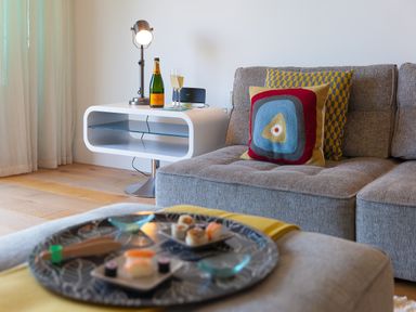 A living room with a gray sofa with patterned cushions a white side table with a lamp champagne bottle and glass and a tray with sushi on a gray ottoman at Crystal Bay in St Ives