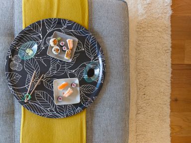 A black tray with white leaf patterns holding two small plates of sushi and dipping sauces on a gray sofa with a yellow cloth at Crystal Bay in St Ives