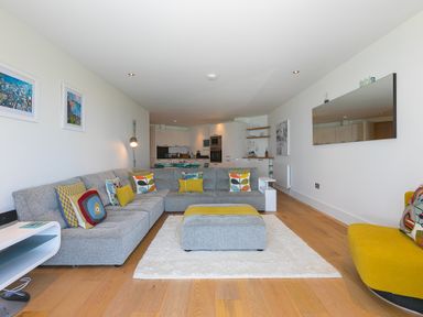 A living room with a gray sectional sofa and yellow armchair adjacent to an open kitchen at Crystal Bay in St Ives
