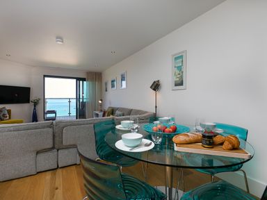 A living room with a gray sofa a round glass dining table with blue chairs and a balcony overlooking the sea at Crystal Bay in St Ives