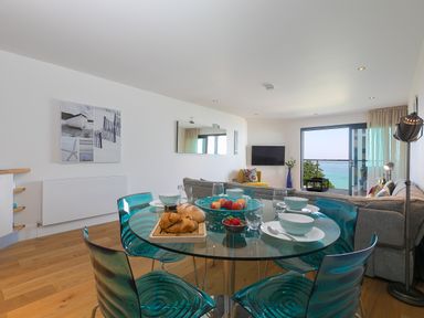 A dining area with a glass table set with bowls and pastries and a living room with a sofa and a television near a balcony with a sea view at Crystal Bay in St Ives
