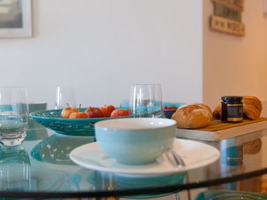 A glass dining table with a bowl on a plate with cutlery a blue bowl of apples bread croissants and a jar on a wooden board at Crystal Bay in St Ives