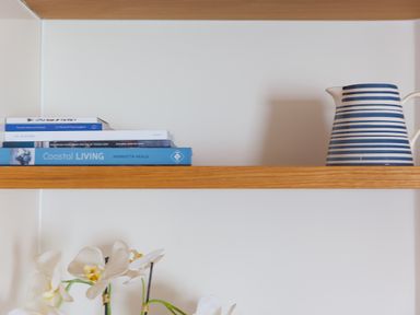 A wooden shelf with stacked books on the left and a striped jug on the right with white flowers below the shelf at Crystal Bay in St Ives