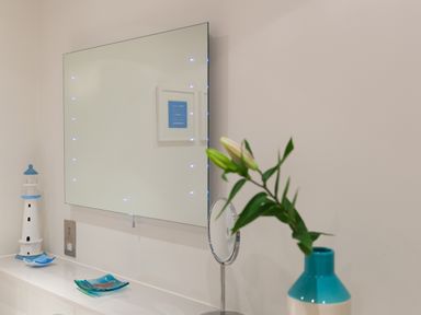 A bathroom counter with a lighthouse decoration a blue vase with green plants a small mirror and a wall-mounted lighted mirror at Crystal Bay in St Ives