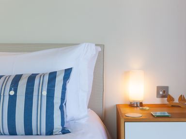 A bed with white sheets and a blue striped pillow next to a wooden bedside table with a lamp and decorative items at Crystal Bay in St Ives