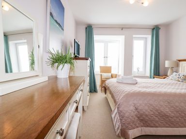 A bedroom with a bed covered with a quilt a dresser with a mirror and a plant a chair by windows with blue curtains at 16 Ocean Heights in Newquay