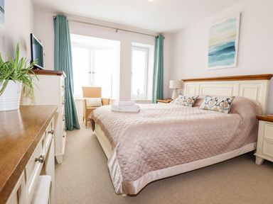 A bedroom with a large bed two windows with blue curtains a chair a dresser and a lamp at 16 Ocean Heights in Newquay