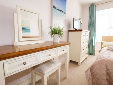 A bedroom with a dressing table mirror a plant a chest of drawers a television a bed and a window with curtains at 16 Ocean Heights in Newquay