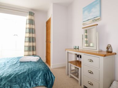 A bedroom with a bed covered by a blue blanket a wooden door striped curtains a dresser with a mirror and a painting above it at 16 Ocean Heights in Newquay