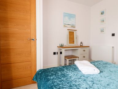 A bedroom with a wooden door a dressing table with a mirror and a stool and a bed with a folded towel on a blue bedspread at 16 Ocean Heights in Newquay