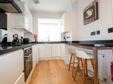 A kitchen with white cabinets black countertops two bar stools under a counter and a cappuccino wall art at 16 Ocean Heights in Newquay