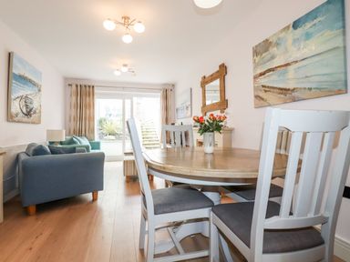A dining area with a wooden round table and white chairs next to a living room with sofas and paintings at 16 Ocean Heights in Newquay