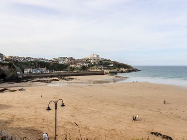 A sandy beach with a few people walking near the shore and houses on a hill by the sea at 16 Ocean Heights in Newquay