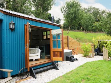 A blue corrugated metal tiny house with open wooden doors showing a white armchair inside a green grassy yard with trees and a golf cart at Little Silver Pheasant in High Bickington