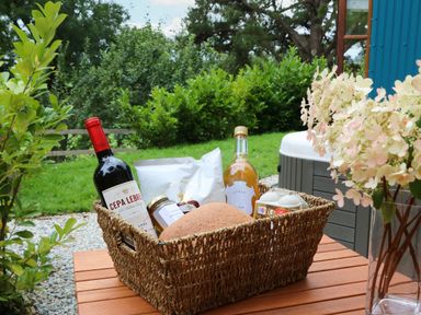 A wicker basket with a bottle of red wine a bottle of drink a loaf of bread jars and a snack bag on a wooden table outdoors with bushes in the background at Little Silver Pheasant in High Bickington