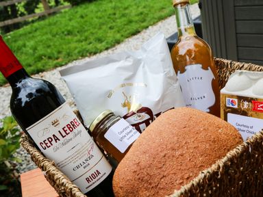 A basket containing a bottle of red wine a loaf of bread a jar of honey a bag of potato chips a bottle of sauce and a carton of eggs at Little Silver Pheasant in High Bickington
