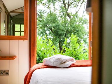A bedroom with a white bedspread and orange blanket with folded robe on top overlooking green bushes and trees at Little Silver Pheasant in High Bickington