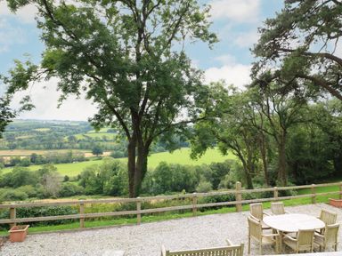 An outdoor patio with wooden table and chairs overlooking trees and green fields at Little Silver Pheasant in High Bickington