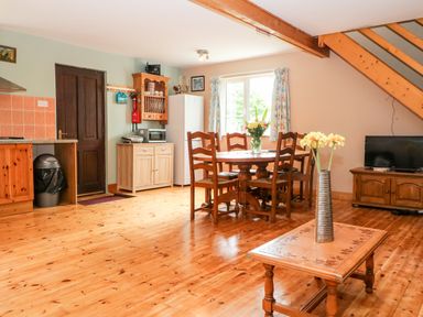 A dining area with wooden table and chairs next to a window a small kitchen area with wooden cabinets a coffee table with flowers a television on a wooden stand under stairs at Teach Chiuin in Coolaheen near Cappoquin County Waterford