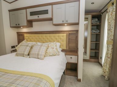 A bedroom with a bed with pillows and a cushioned headboard with overhead cabinets and an open wardrobe with shelves at 22 Sherwood South Lakeland Leisure Village near Warton