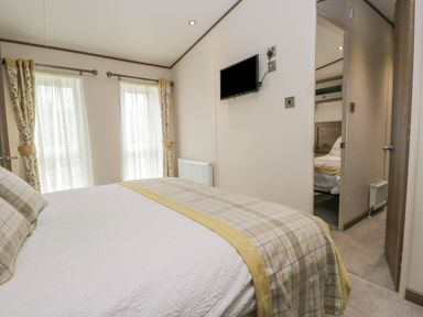 A bedroom with a bed covered in a white and plaid yellow blanket near two windows with floral curtains and a wall-mounted TV at 22 Sherwood in South Lakeland Leisure Village near Warton