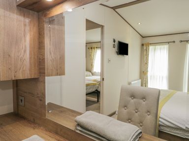 A bedroom with a bed a wall-mounted tv a window with curtains and a dressing table with a chair at 22 Sherwood South Lakeland Leisure Village near Warton